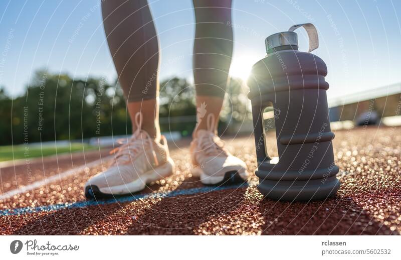 Nahaufnahme einer großen Wasserflasche auf einer Laufstrecke mit Frauenbeinen im Hintergrund Laufbahn Fitness Hydratation Sportbekleidung Athlet Training