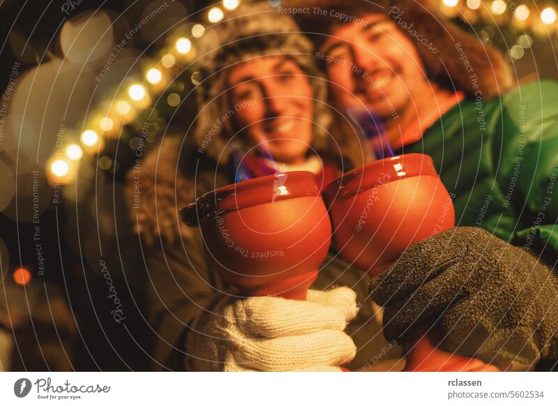 Lächelndes Paar auf einem Weihnachtsmarkt, rote Tassen mit blauen Flammen aus Feuerzangenbowle in der Hand, dahinter festliche Lichter. Tourist Glühwein