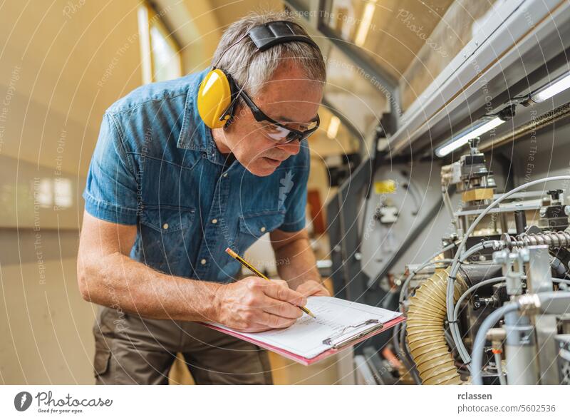 Techniker mit Gehörschutz beim Schreiben auf einem Klemmbrett in einer industriellen Umgebung schreibend Zwischenablage Maschine Werkstatt Besichtigung