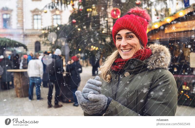 Lächelnde Frau in roter Mütze und Schal mit Schneeflocken auf einem festlichen Weihnachtsmarkt zur Winterzeit in Deutschland, mit Platz für Ihren individuellen Text.