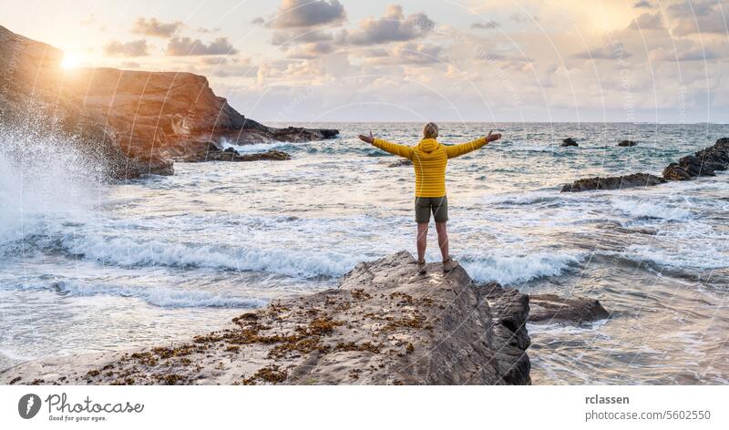 Person in gelber Jacke steht auf einem Felsen am Meer mit weit geöffneten Armen bei Sonnenuntergang Fuerteventura Freiheit MEER Natur gelbe Jacke Küstenlinie