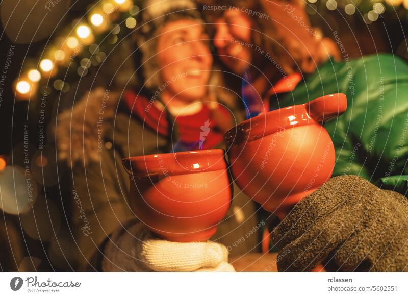 Lachendes Paar, das auf einem Weihnachtsmarkt Feuerzangenbowle genießt und dampfende rote Becher mit blauen Flammen in der Hand hält. Tourist Glühwein