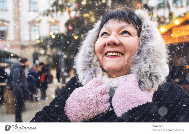 Ältere Frau in festlicher Kleidung lächelt auf einem winterlichen Weihnachtsmarkt, während Schnee fällt Glück Frohe Weihnachten Kakao Handschuhe traditionell