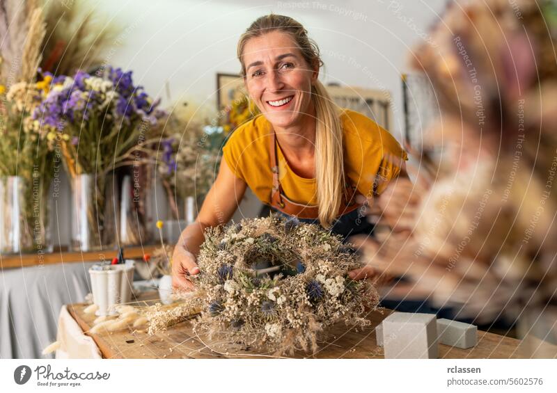 Fröhliche Floristin, die in einer hellen Blumenwerkstatt einen Kranz aus Trockenblumen hält und bearbeitet freudig Blumenhändlerin mit Kranz in der Hand