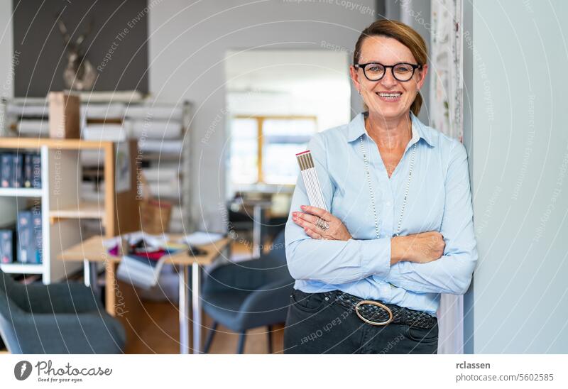professionelle lächelnde Schneiderin mit geflochtenem Haar, die eine Brille und ein blau gestreiftes Hemd trägt, steht in einem modernen Arbeitsraum und hält ein Lineal.