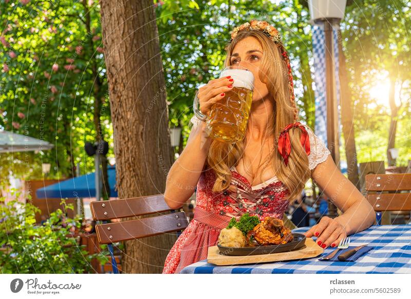Frau im Dirndl im bayerischen Biergarten sitzend und Bier trinkend mit traditioneller bayerischer Küche mit Schweinshaxe, gebratener Schweinshaxe auf einem Tisch