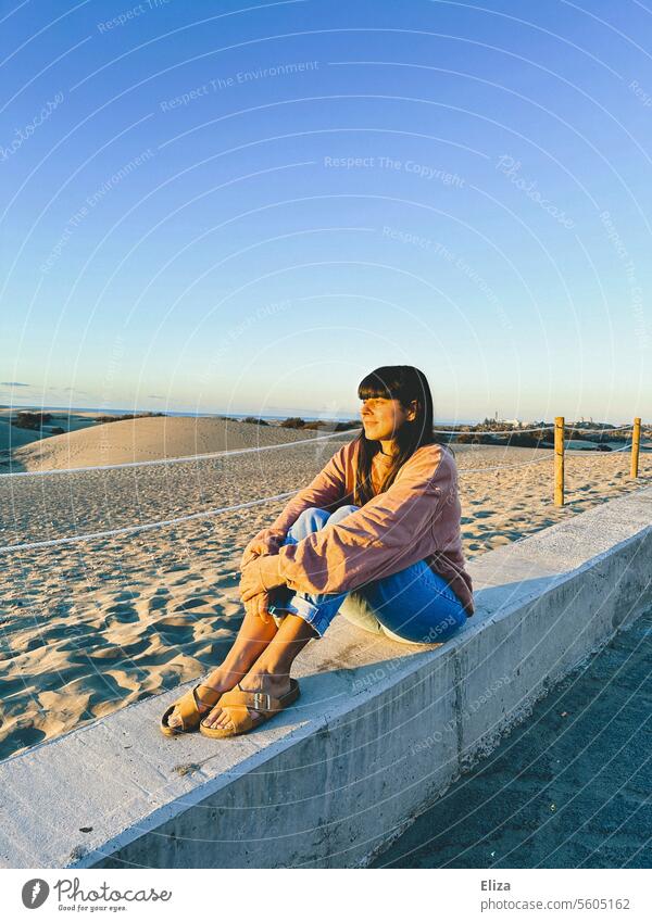 Frau sitzt auf einer Steinmauer vor den Dünen von Maspalomas und blickt in die Sonne Gran Canaria Gesicht Sonnenschein Urlaub sitzen Sand Natur Sommer Himmel