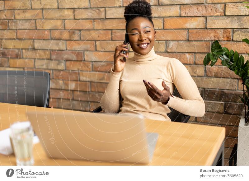 Eine junge afroamerikanische Geschäftsfrau benutzt ein Mobiltelefon, während sie an der Backsteinmauer in einem Büro im Industriestil sitzt. Erwachsener