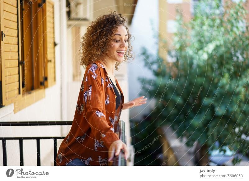 Fröhliche junge brünette Frau mit lockigem Haar steht auf dem Balkon eines modernen Gebäudes Lächeln heiter Stadt Glück Inhalt froh lässig positiv Haus angenehm