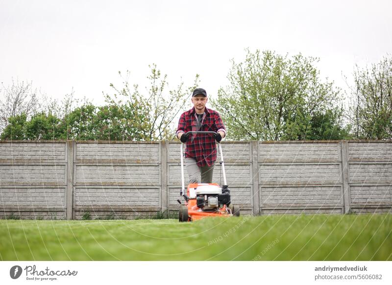 Blick auf Mann in Freizeitkleidung mäht Rasen mit Rasenmäher im Hinterhof seines Hauses. Der Ehemann kümmert sich um den Garten an einem bewölkten Frühlingstag. Moderne Benzin-Gartengeräte. Landschaftsbau Arbeit