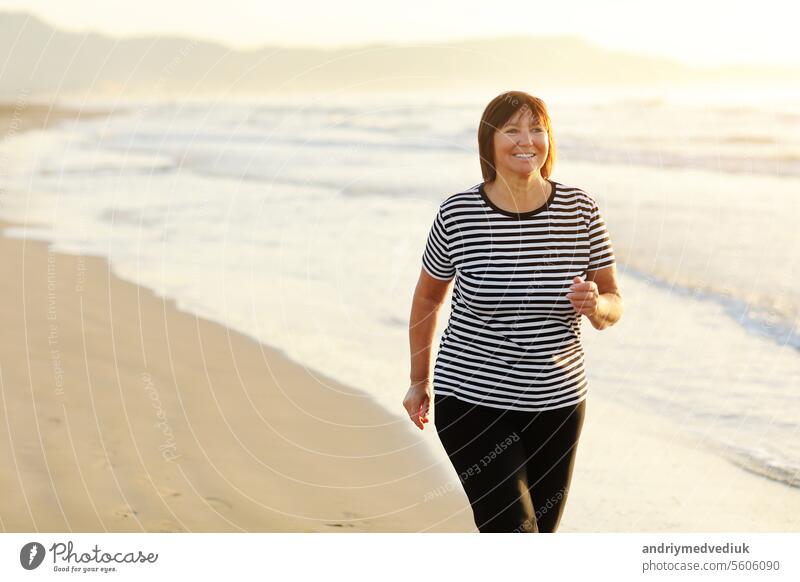Lächelnde Frau mittleren Alters läuft am Strand bei Sonnenaufgang. 40s oder 50s attraktive reife Dame in Sportkleidung tun Jogging Training genießen Fitness und gesunden Lebensstil am schönen Meer Landschaft.