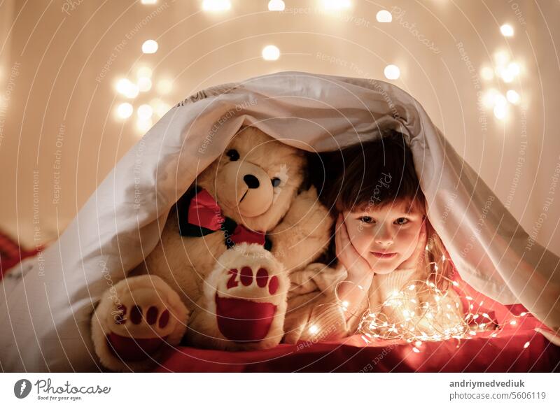kleine niedliche lächelnde Baby-Mädchen mit weißen Teddybär späht aus unter einer Decke auf einem Bett zu Hause mit Beleuchtung Girlanden bei Dunkelheit. Kind schaut in die Kamera. Fairytale Kindheit mit Wunder Konzept