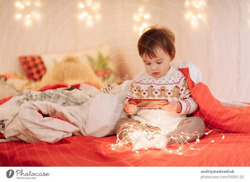entzückende kleine Mädchen in Wurmkleidung mit Winterschmuck haben Spaß, kommunizieren und teilen Geheimnisse auf einem Bett unter einer Decke in einem gemütlichen Schlafzimmer dekoriert Girlanden, Neujahrs-Familienurlaub
