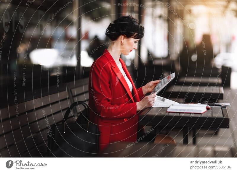 Eine attraktive Geschäftsfrau in Corporate Outfit roten Blazer hält Morgenzeitung beim Sitzen auf Cafe Terrasse. Manager liest die neuesten Weltnachrichten während einer Kaffeepause bei der Arbeit