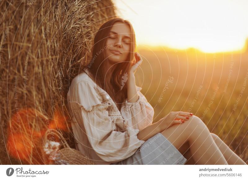 Junge Frau im schönen Licht des Sommers Sonnenuntergang in einem Feld sitzt in der Nähe der Strohballen. schöne romantische Mädchen mit langen Haaren im Freien im Feld