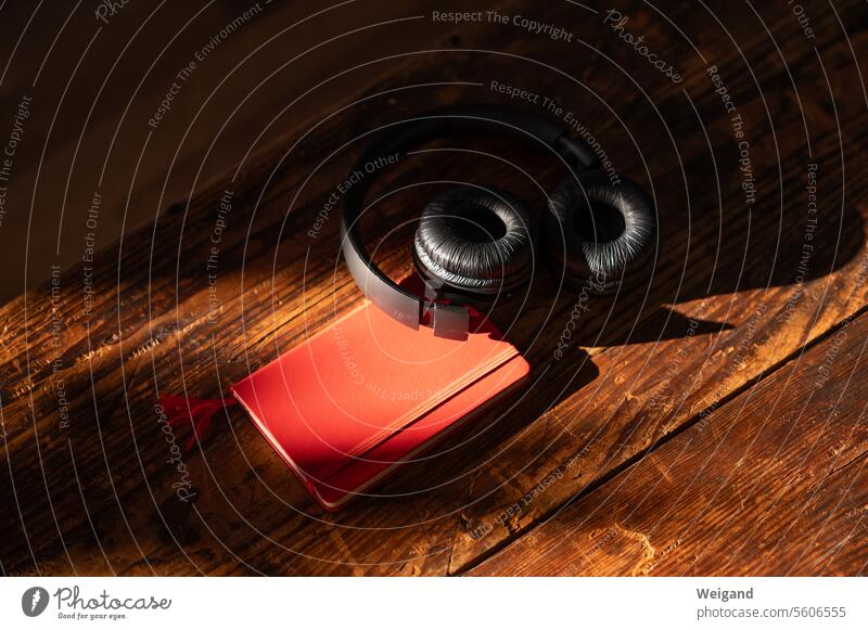Schwarze Kopfhörer liegen in atmosphärischer Stimmung auf einem vom Sonnenlicht erfassten roten Notizbuch auf dunkelbraun gemaserten Holzdielen, bereit, der Musik zu lauschen und die Gedanken nieder zu schreiben