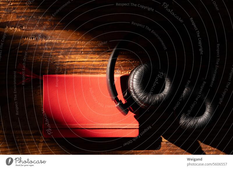 Schwarze Kopfhörer liegen in atmosphärischer Stimmung auf einem vom Sonnenlicht erfassten roten Notizbuch auf einem dunkel gemaserten Holzuntergrund, bereit, der Musik zu lauschen und die Gedanken nieder zu schreiben