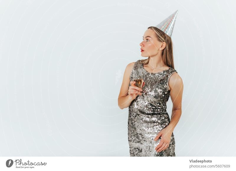 Junge kaukasische blonde Frau mit Weinglas in silbernem Glitzerkleid und Partyhut feiert auf weißem Hintergrund. Champagne Glas Party-Kappe glänzend Kleid