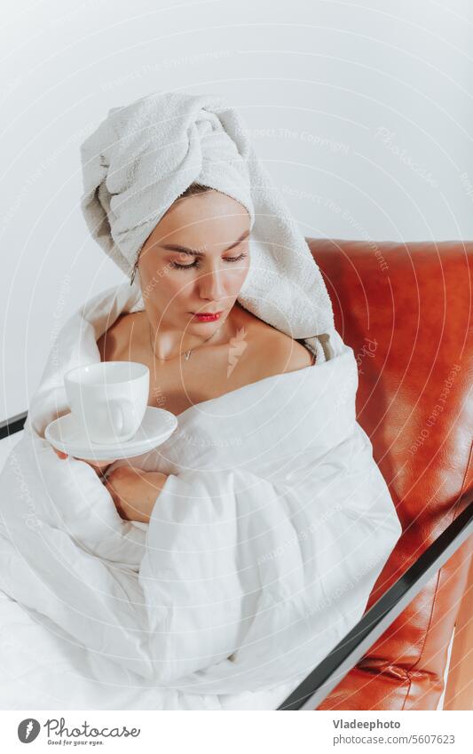 Frau trinkt Kaffee oder Tee, eingewickelt in eine weiße Decke und ein Handtuch um den Kopf. Weißer Hintergrund Morgen heimwärts Lifestyle Bademantel Streu