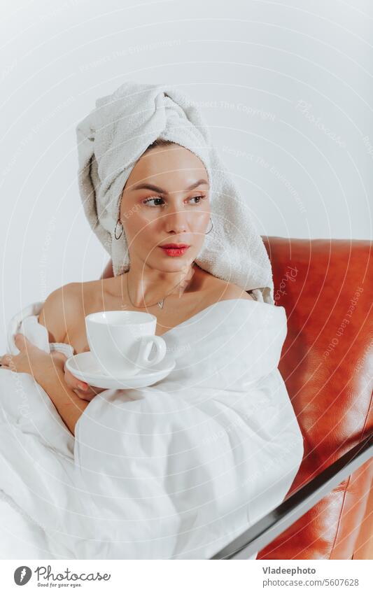 Frau trinkt Kaffee oder Tee, eingewickelt in eine weiße Decke und ein Handtuch um den Kopf. Weißer Hintergrund Morgen heimwärts Lifestyle Bademantel Streu