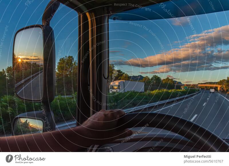 Blick aus dem Fahrersitz eines Lastwagens auf eine Autobahn in der Morgendämmerung und die im Rückspiegel reflektierte Sonne. Hand im Inneren Lenkrad