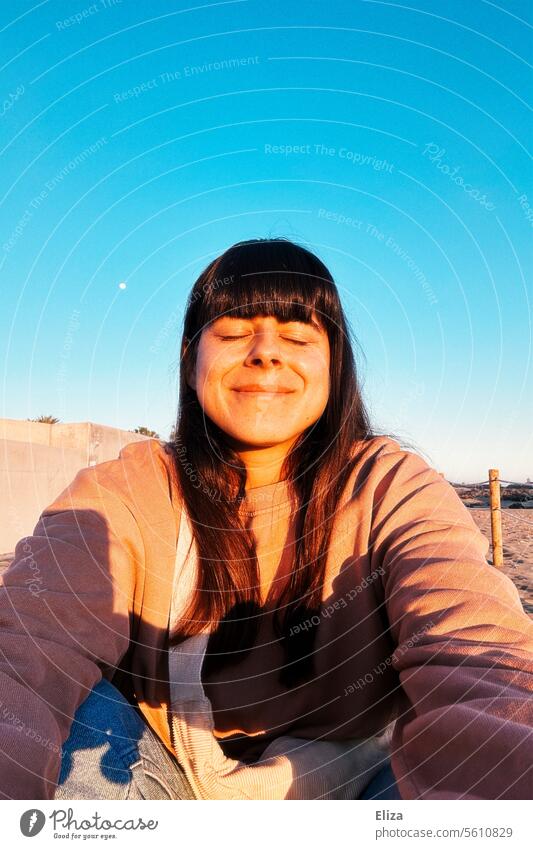 Selfie einer zufrieden lächelnden Frau mit geschlossenen Augen in der Morgensonne vor blauem Himmel geschlossene Augen Sonne Zufriedenheit Lächeln Porträt