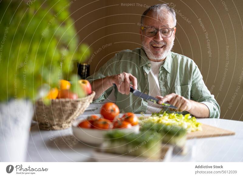 Fröhlicher älterer Mann mit Brille schneidet Salat in Stücke mit einem Messer auf einem hölzernen Schneidebrett, während er einen Salat zu Hause zubereitet