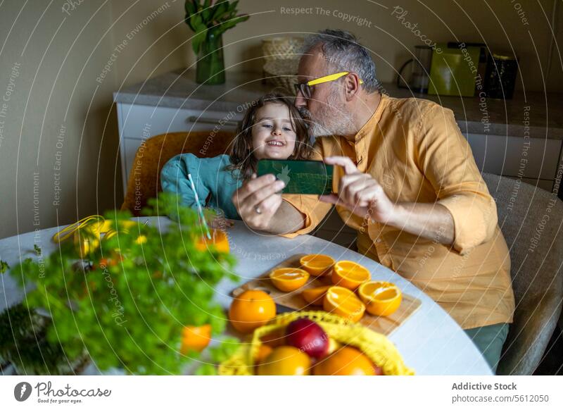 Hoher Winkel Blick auf ältere Großvater küssen auf glückliche Enkelin die Stirn während der Aufnahme von Selfie über Smartphone am Esstisch zu Hause Mädchen