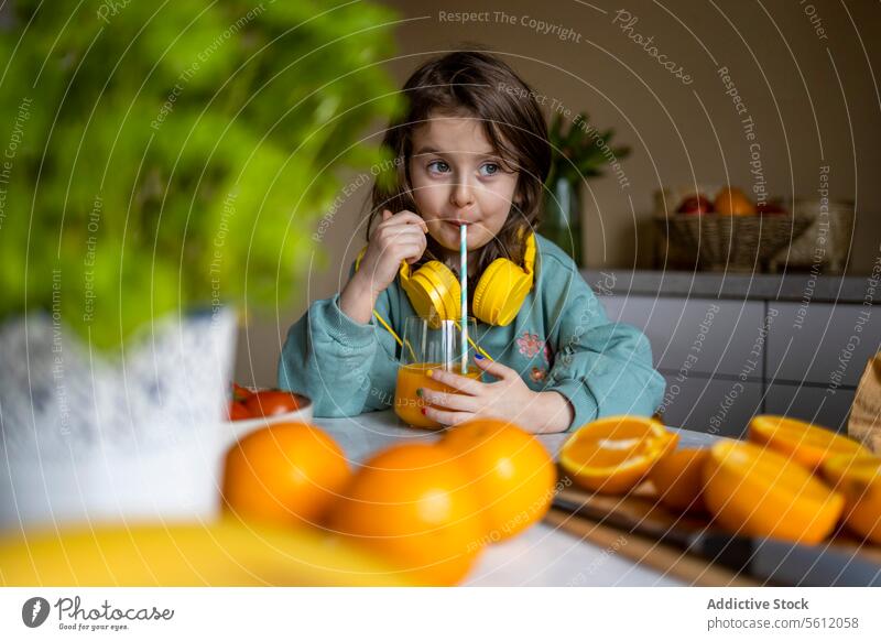 Lächelndes bezauberndes Mädchen mit Kopfhörern im Nacken, das wegschaut, während es Orangensaft auf einem Tisch zu Hause trinkt Saft Glas Stroh heimwärts