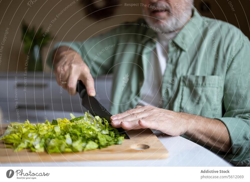 Anonymer älterer Mann schneidet Salat mit einem Messer auf einem hölzernen Schneidebrett in Stücke, während er einen Salat zu Hause zubereitet Senior Ernte
