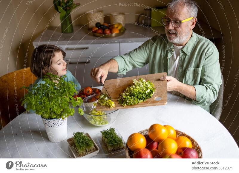 Mann bereitet Gemüsesalat mit Kind am Tisch zu Großvater Enkelin Mädchen Senior Frucht Pflanze Kraut Schneidebrett Schalen & Schüsseln Messer Lernen älter Salat