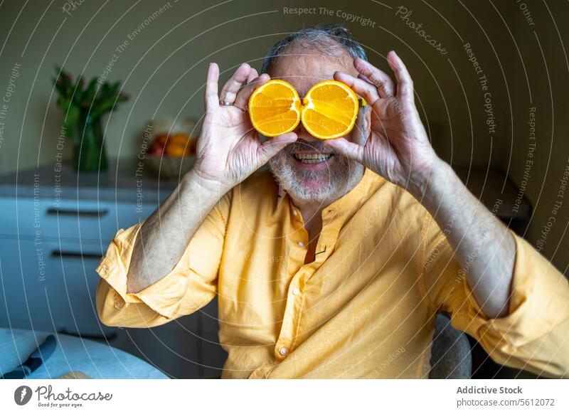 Älterer Mann bedeckt zu Hause seine Augen mit Orangenscheiben Senior orange Lächeln deckend Scheibe halbiert heiter Küche Sitzen Freizeitkleidung Lifestyle