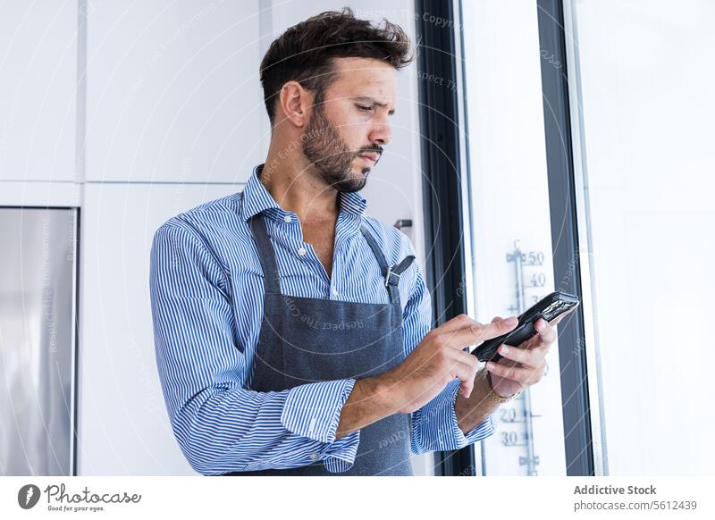 Seitenansicht eines konzentriert wirkenden Mannes in Schürze, der bei Tageslicht in der Nähe eines Fensters auf seinem Smartphone surft Küchenchef benutzend