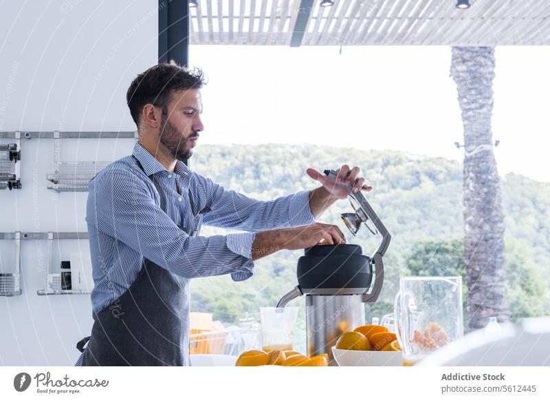 Seitenansicht eines bärtigen Mannes in Schürze, der in der Nähe eines Tisches mit einer Orangenpresse in einer modernen Küche steht und Orangensaft zubereitet