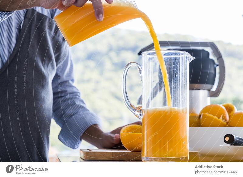 Unbekannter Koch in Schürze steht am Küchentisch und gießt Orangensaft in einen Krug in einer modernen Küche Mann Küchenchef eingießen Saft Frühstück drücken