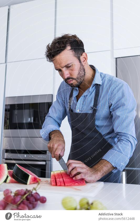 Privater bärtiger Koch in Schürze steht am Tisch und schneidet Wassermelone in der Küche Mann Küchenchef geschnitten Scheibe Messer gesunde Ernährung Vitamin