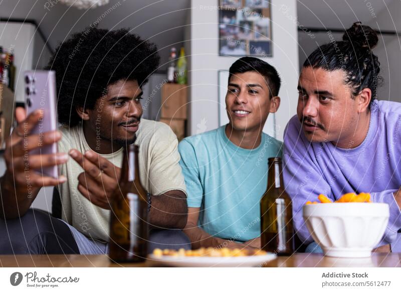Männliche Freunde sehen ein Foto auf dem Smartphone zu Hause Lächeln Afrohaar Freizeit Zusammensein Wohnzimmer Freundschaft Wochenende Mobile Telefon Bier