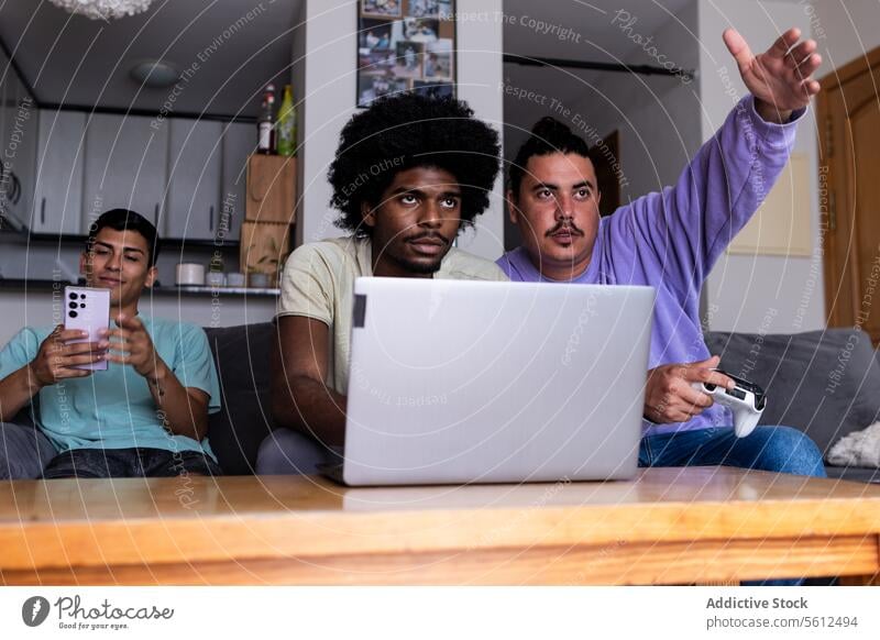 Multirassische männliche Freunde, die zu Hause Technologie nutzen Smartphone Laptop Konsole Zeigen Wegsehen Computer Tisch Wohnzimmer Wochenende vielfältig
