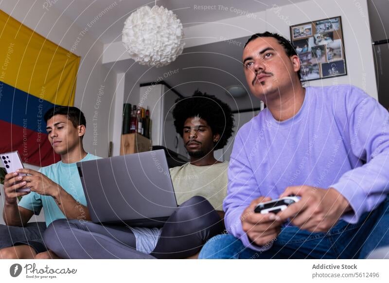 Multirassische männliche Freunde, die zu Hause Technologie nutzen Smartphone Laptop Konsole Zeigen Blick auf den Bildschirm Computer Tisch Wohnzimmer Wochenende