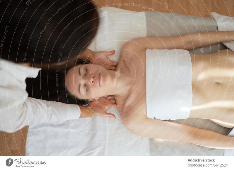 Draufsicht auf eine in ein weißes Handtuch eingewickelte Kundin mit geschlossenen Augen, die auf dem Bett liegt und eine Nackentherapie-Massage von einem nicht erkennbaren Therapeuten erhält