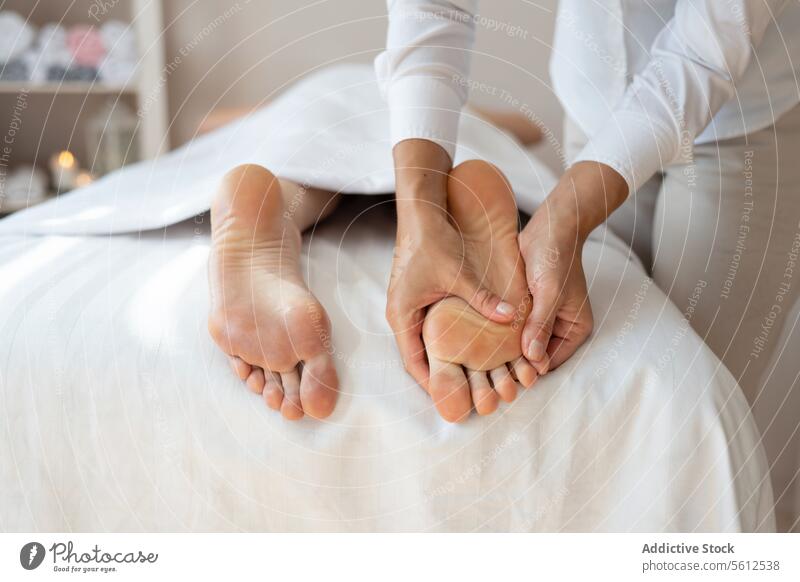 Die Hände einer anonymen weiblichen Therapeutin massieren den Fuß einer auf dem Bett liegenden Kundin, um Schmerzen durch sanften Druck auf die Druckpunkte in einem Wellness-Salon zu lindern.