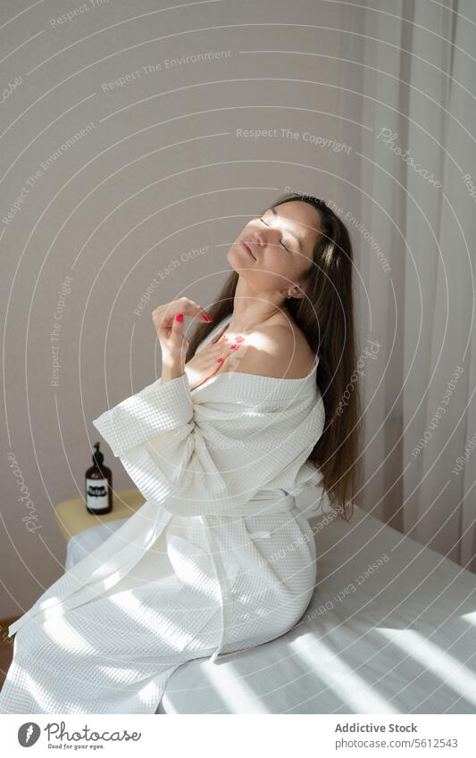 Positive, ruhige, erwachsene Frau in weißem Bademantel mit geschlossenen Augen, die ihren Körper berührt und sich entspannt, während sie auf dem Bett sitzt, nachdem sie im Wellnessbereich war