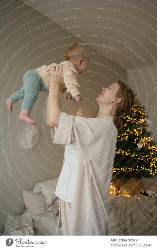 Junge Mutter hebt Baby mit Weihnachtsbaum im Hintergrund Weihnachten Baum heimwärts Freude Angebot heben warm Licht gemütlich im Innenbereich Feier festlich