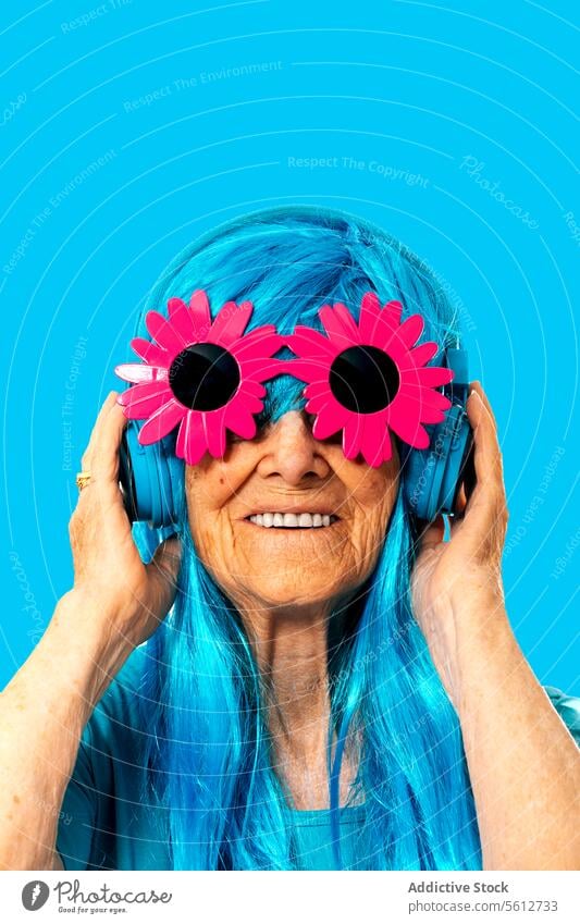 Porträt einer glücklichen älteren Frau mit blauer Perücke, Kopfhörern und Sonnenbrille mit Blumenrahmen vor blauem Hintergrund Hautfalten Glück Vorschein