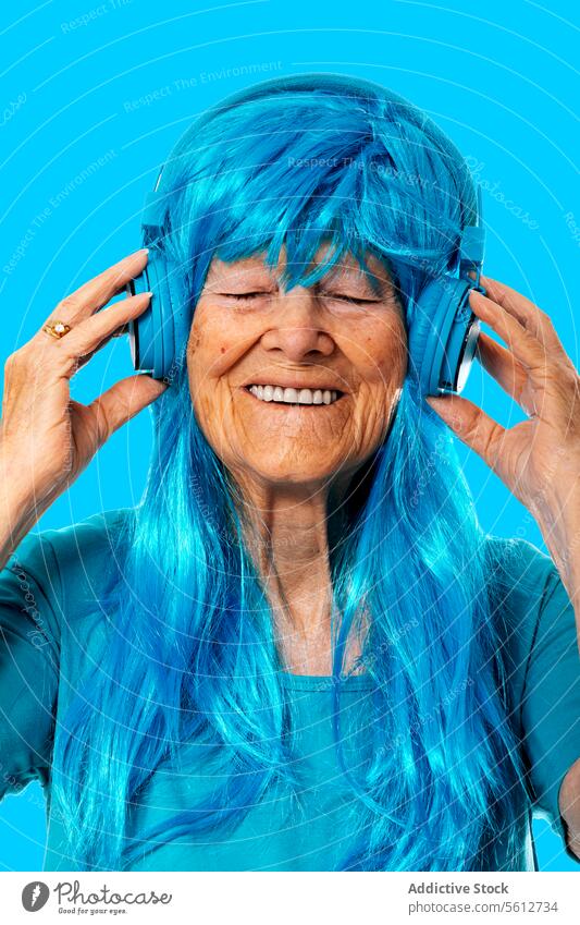 Fröhliche ältere Frau, die mit geschlossenen Augen auf blauem Hintergrund über Kopfhörer Musik hört Porträt zuhören genießen Gesang Perücke Farbe Streichholz