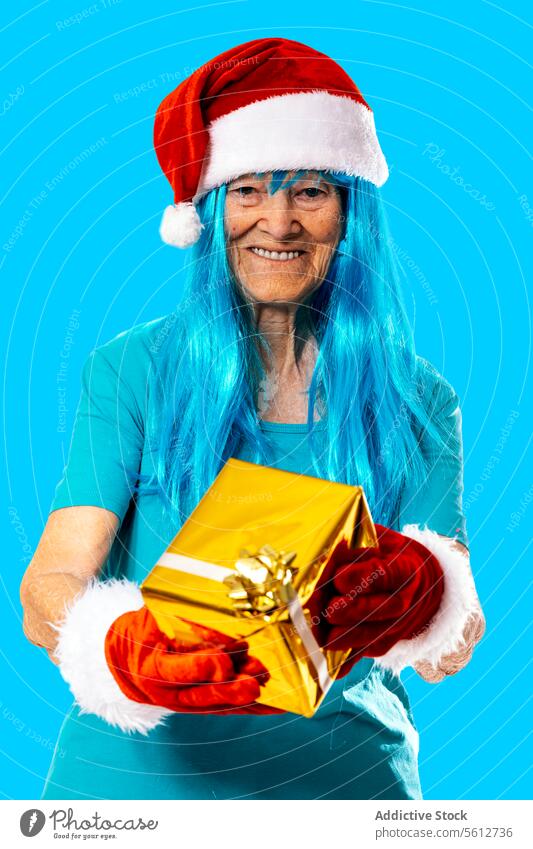 Fröhliche ältere Frau mit blauer Perücke, Weihnachtsmannmütze und Handschuhen, die der Kamera ein Geschenk auf blauem Hintergrund überreicht Weihnachten geben