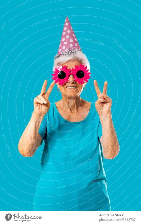 Lustige ältere Frau mit lustiger rosa Sonnenbrille und Geburtstagskappe zeigt zwei Finger im Studio Verschlussdeckel zeigen gestikulieren Frieden feiern Lächeln