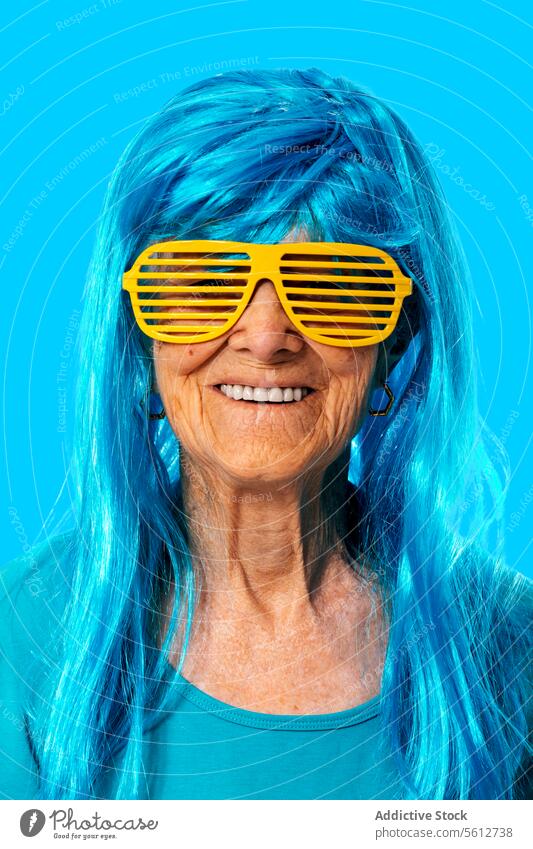Lächelnde ältere Frau mit Sonnenbrille und blauer Perücke schaut in die Kamera, während sie auf blauem Hintergrund steht Porträt Streifen in den Ruhestand gehen