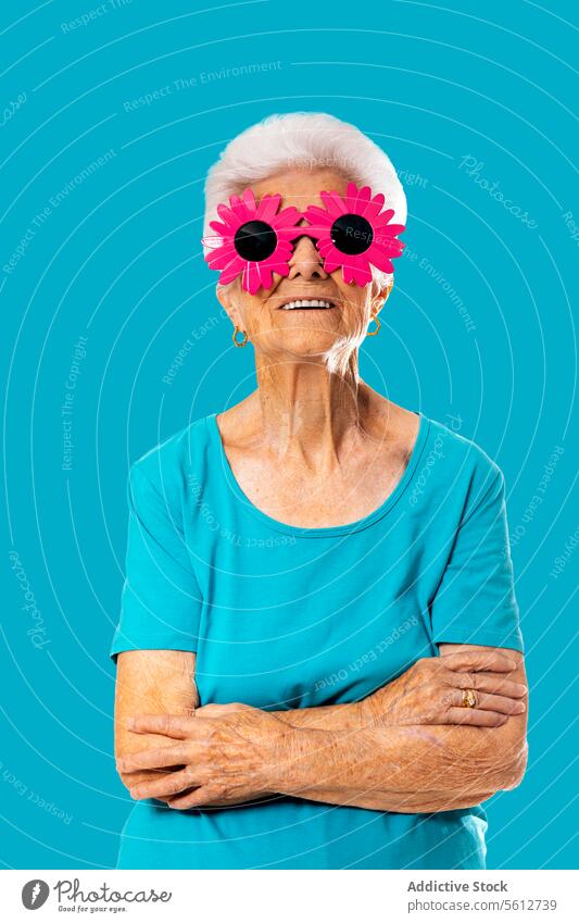 Fröhliche ältere Frau, die eine lustige rosa Sonnenbrille trägt und mit verschränkten Armen auf blauem Hintergrund steht die Arme verschränkt kindisch