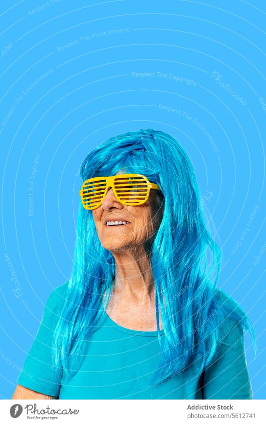 Lächelnde ältere Frau mit Sonnenbrille und blauer Perücke schaut weg, während sie auf blauem Hintergrund steht Porträt Streifen in den Ruhestand gehen Rentnerin